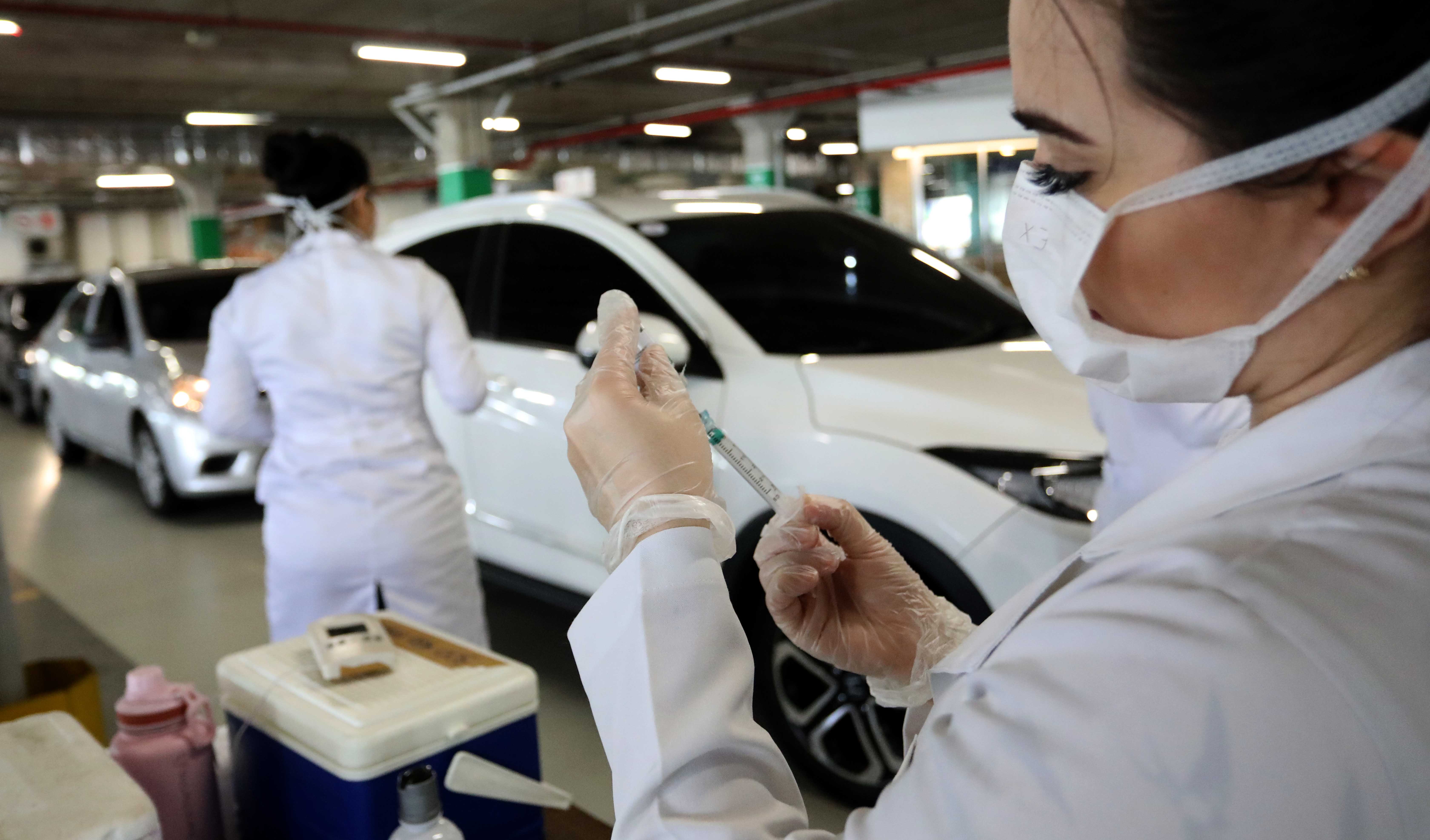 enfermeira manuseando seringa e carros e outras enfermeiras ao fundo em estacionamento de shopping